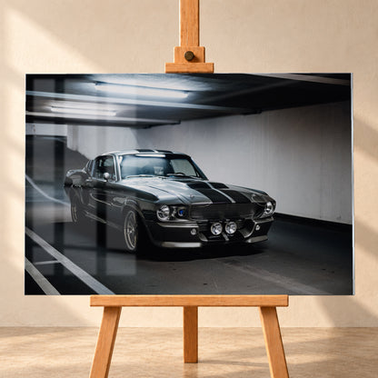 Eleanor mit dunkler Karosserie in frontaler Dreiviertelansicht in einer Beton-Parkgarage; sichtbar sind Längsstreifen, Rundscheinwerfer, zwei zentrale Zusatzleuchten, Fahrbahnmarkierungen und helle Deckenröhren; Visualisierung als Acrylglas-Wandbild, frontal auf einer Holzstaffelei.