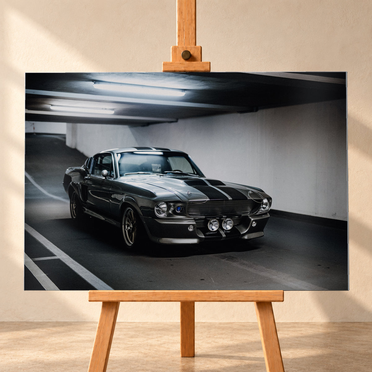 Eleanor mit dunkler Karosserie in frontaler Dreiviertelansicht in einer Beton-Parkgarage; sichtbar sind Längsstreifen, Rundscheinwerfer, zwei zentrale Zusatzleuchten, Fahrbahnmarkierungen und helle Deckenröhren; Visualisierung als Alu gebürstet-Wandbild, frontal auf einer Holzstaffelei.