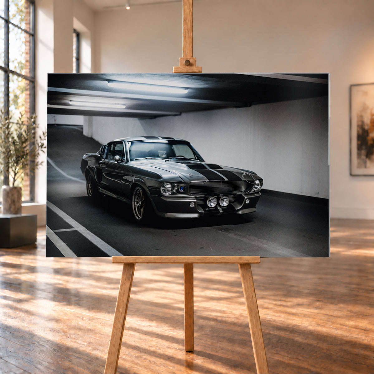 Eleanor mit dunkler Karosserie in frontaler Dreiviertelansicht in einer Beton-Parkgarage; sichtbar sind Längsstreifen, Rundscheinwerfer, zwei zentrale Zusatzleuchten, Fahrbahnmarkierungen und helle Deckenröhren; Visualisierung als Alu gebürstet-Wandbild, auf einer Holzstaffelei in einem hellen Galerieraum.