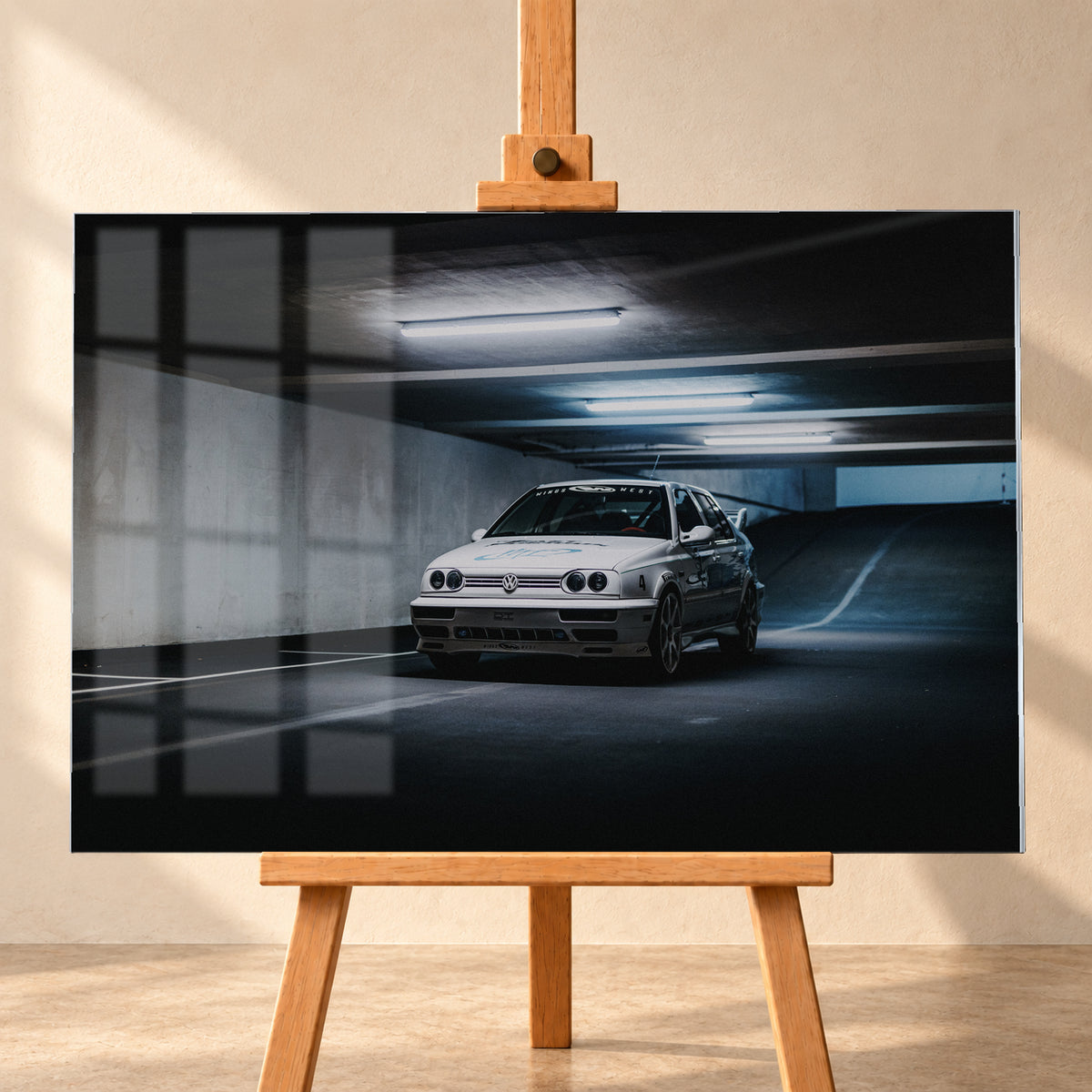 Weißer Volkswagen Jetta in tiefer Dreiviertelansicht von vorn in einer dunklen Beton-Parkgarage. Kühle Deckenleuchten, markierte Stellflächen und eine ansteigende Rampe rahmen das Fahrzeug mit großen Rädern, dunklen Seitengrafiken und Heckflügel; Visualisierung als Acrylglas-Wandbild, frontal auf einer Holzstaffelei.