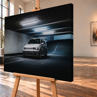 Weißer Volkswagen Jetta in tiefer Dreiviertelansicht von vorn in einer dunklen Beton-Parkgarage. Kühle Deckenleuchten, markierte Stellflächen und eine ansteigende Rampe rahmen das Fahrzeug mit großen Rädern, dunklen Seitengrafiken und Heckflügel; Visualisierung als Leinwand-Wandbild, schräg von der Seite auf einer Holzstaffelei.