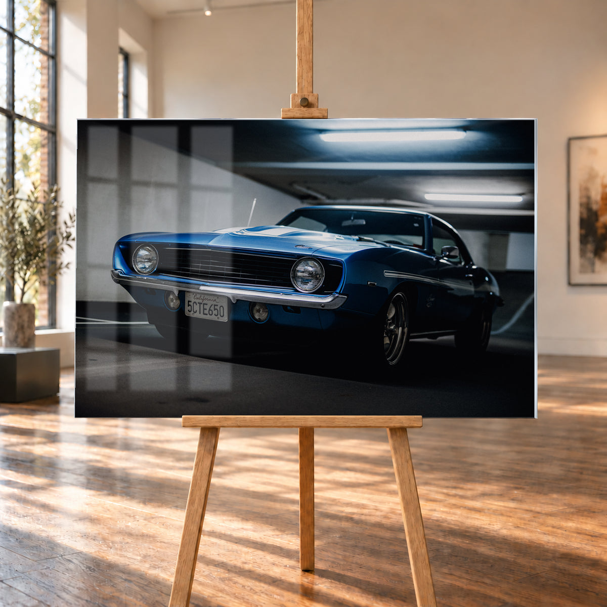 Blauer Yenko Camaro in tiefer Front-Dreiviertelansicht in einer dunklen Parkgarage; sichtbar sind runde Scheinwerfer, schwarzer Kühlergrill, Chromstoßstange, helle Streifen und leuchtende Deckenröhren; Visualisierung als Acrylglas-Wandbild, auf einer Holzstaffelei in einem hellen Galerieraum.