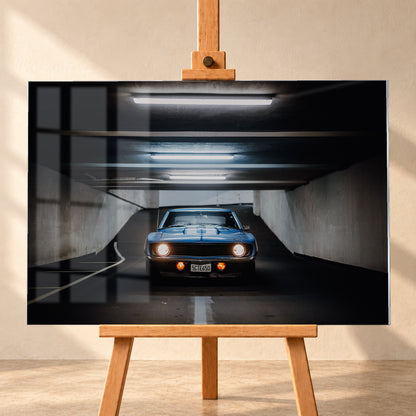 Frontansicht eines blauen Chevrolet Camaro in einer Beton-Tiefgarage, mittig auf dunkler Fahrbahn unter hellen Deckenröhren, mit leuchtenden runden Scheinwerfern, orangefarbenen Zusatzleuchten und weißen Fahrbahnmarkierungen; Visualisierung als Acrylglas-Wandbild, frontal auf einer Holzstaffelei.
