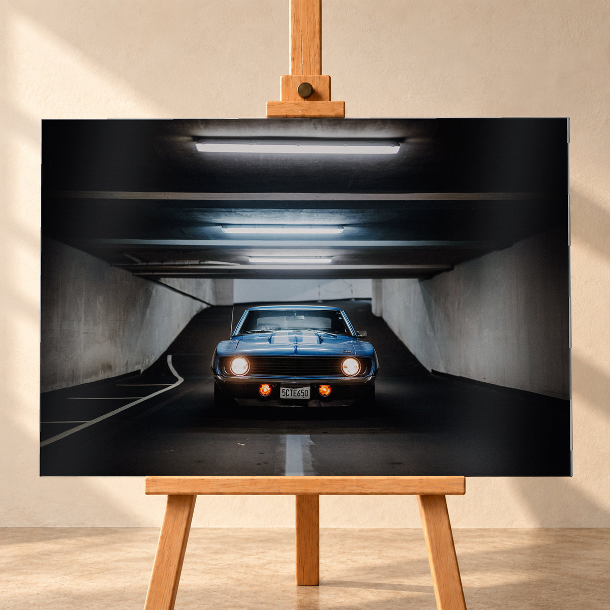 Frontansicht eines blauen Chevrolet Camaro in einer Beton-Tiefgarage, mittig auf dunkler Fahrbahn unter hellen Deckenröhren, mit leuchtenden runden Scheinwerfern, orangefarbenen Zusatzleuchten und weißen Fahrbahnmarkierungen; Visualisierung als Alu gebürstet-Wandbild, frontal auf einer Holzstaffelei.