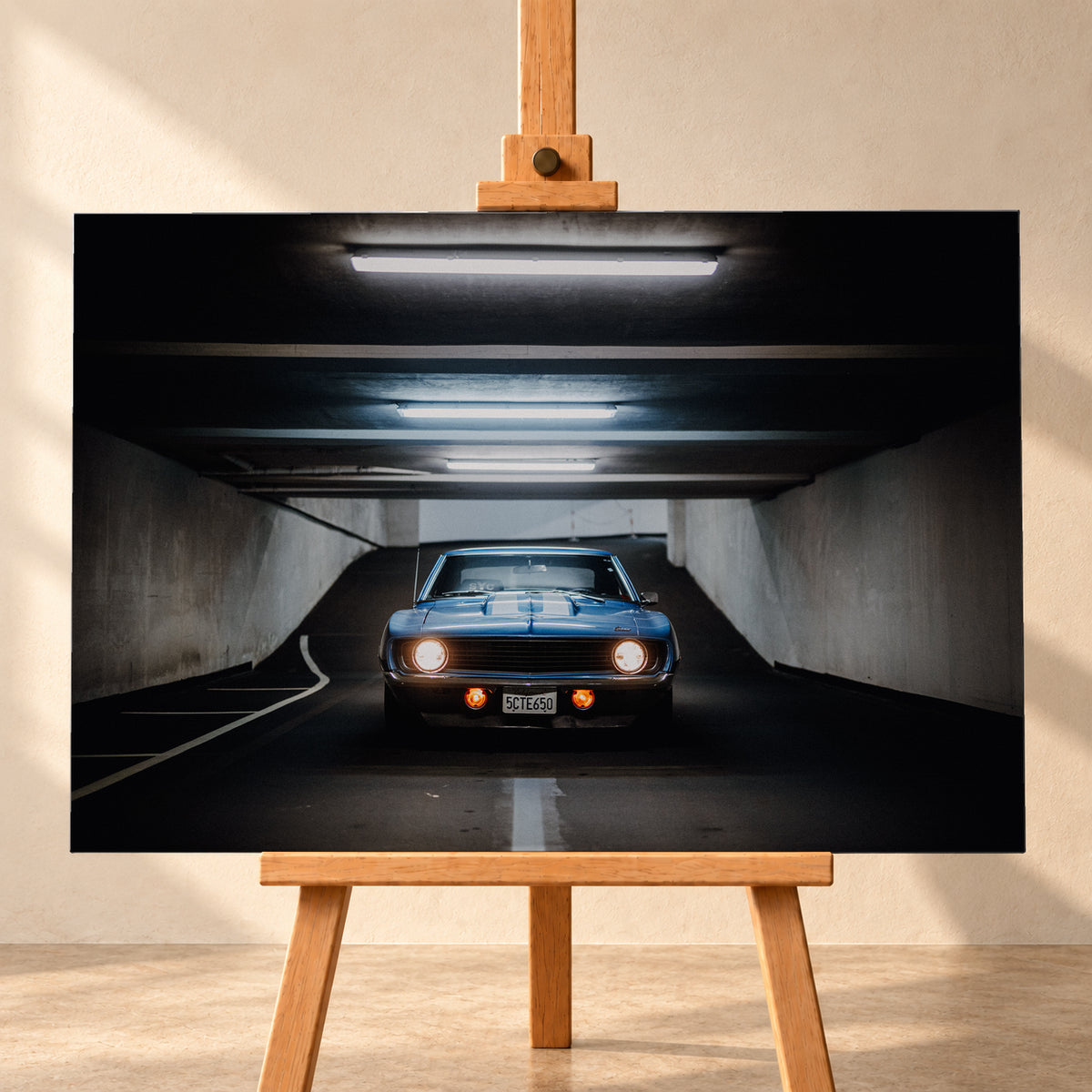 Frontansicht eines blauen Chevrolet Camaro in einer Beton-Tiefgarage, mittig auf dunkler Fahrbahn unter hellen Deckenröhren, mit leuchtenden runden Scheinwerfern, orangefarbenen Zusatzleuchten und weißen Fahrbahnmarkierungen; Visualisierung als Leinwand-Wandbild, frontal auf einer Holzstaffelei.