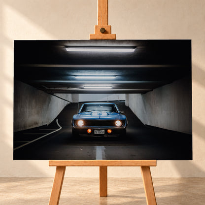 Frontansicht eines blauen Chevrolet Camaro in einer Beton-Tiefgarage, mittig auf dunkler Fahrbahn unter hellen Deckenröhren, mit leuchtenden runden Scheinwerfern, orangefarbenen Zusatzleuchten und weißen Fahrbahnmarkierungen; Visualisierung als Leinwand-Wandbild, frontal auf einer Holzstaffelei.
