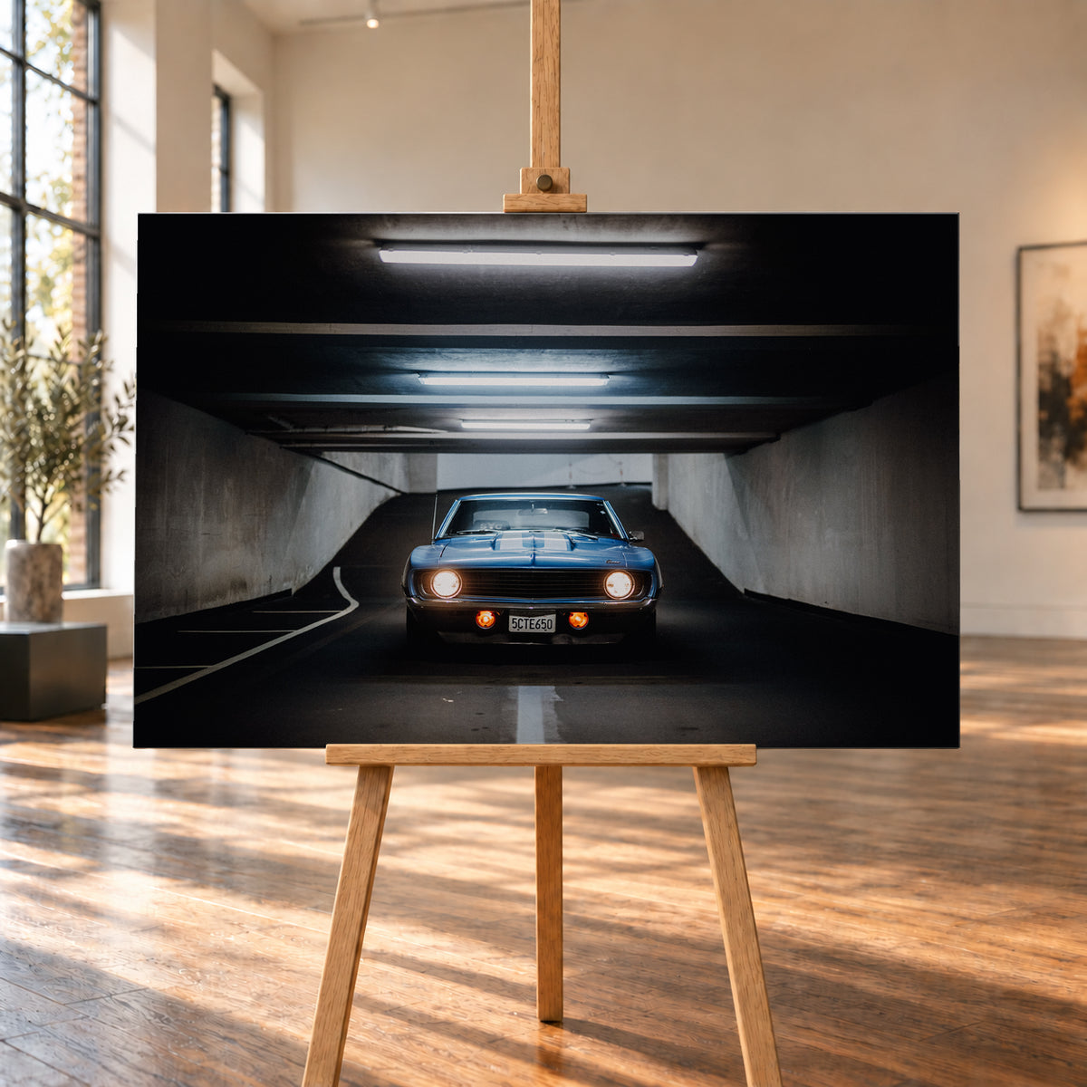 Frontansicht eines blauen Chevrolet Camaro in einer Beton-Tiefgarage, mittig auf dunkler Fahrbahn unter hellen Deckenröhren, mit leuchtenden runden Scheinwerfern, orangefarbenen Zusatzleuchten und weißen Fahrbahnmarkierungen; Visualisierung als Leinwand-Wandbild, auf einer Holzstaffelei in einem hellen Galerieraum.