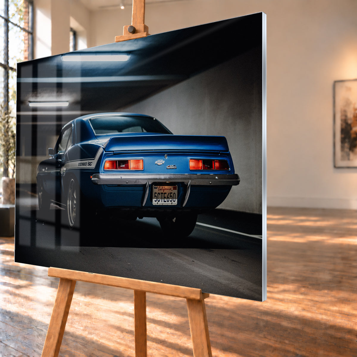Blauer Chevrolet Camaro Yenko in tiefer Heck-Dreiviertelansicht in einer dunklen Parkgarage; rote Rücklichter, Heckspoiler, YENKO/SC-Seitenstreifen und kühle Deckenleuchten sind sichtbar; Visualisierung als Acrylglas-Wandbild, schräg von der Seite auf einer Holzstaffelei.