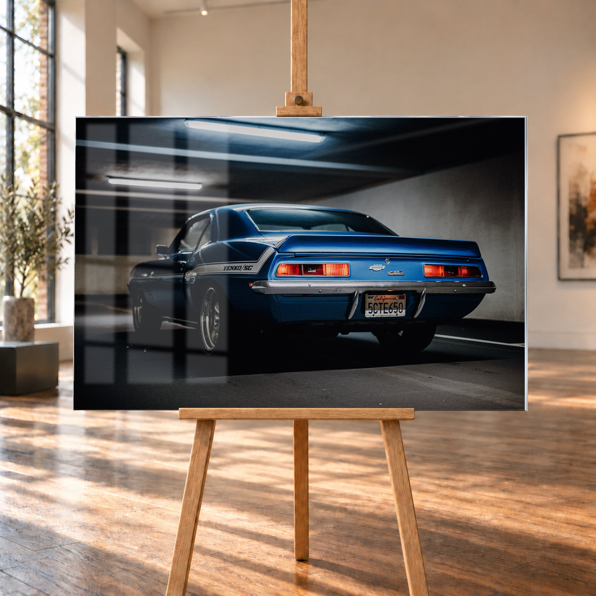 Blauer Chevrolet Camaro Yenko in tiefer Heck-Dreiviertelansicht in einer dunklen Parkgarage; rote Rücklichter, Heckspoiler, YENKO/SC-Seitenstreifen und kühle Deckenleuchten sind sichtbar; Visualisierung als Acrylglas-Wandbild, auf einer Holzstaffelei in einem hellen Galerieraum.