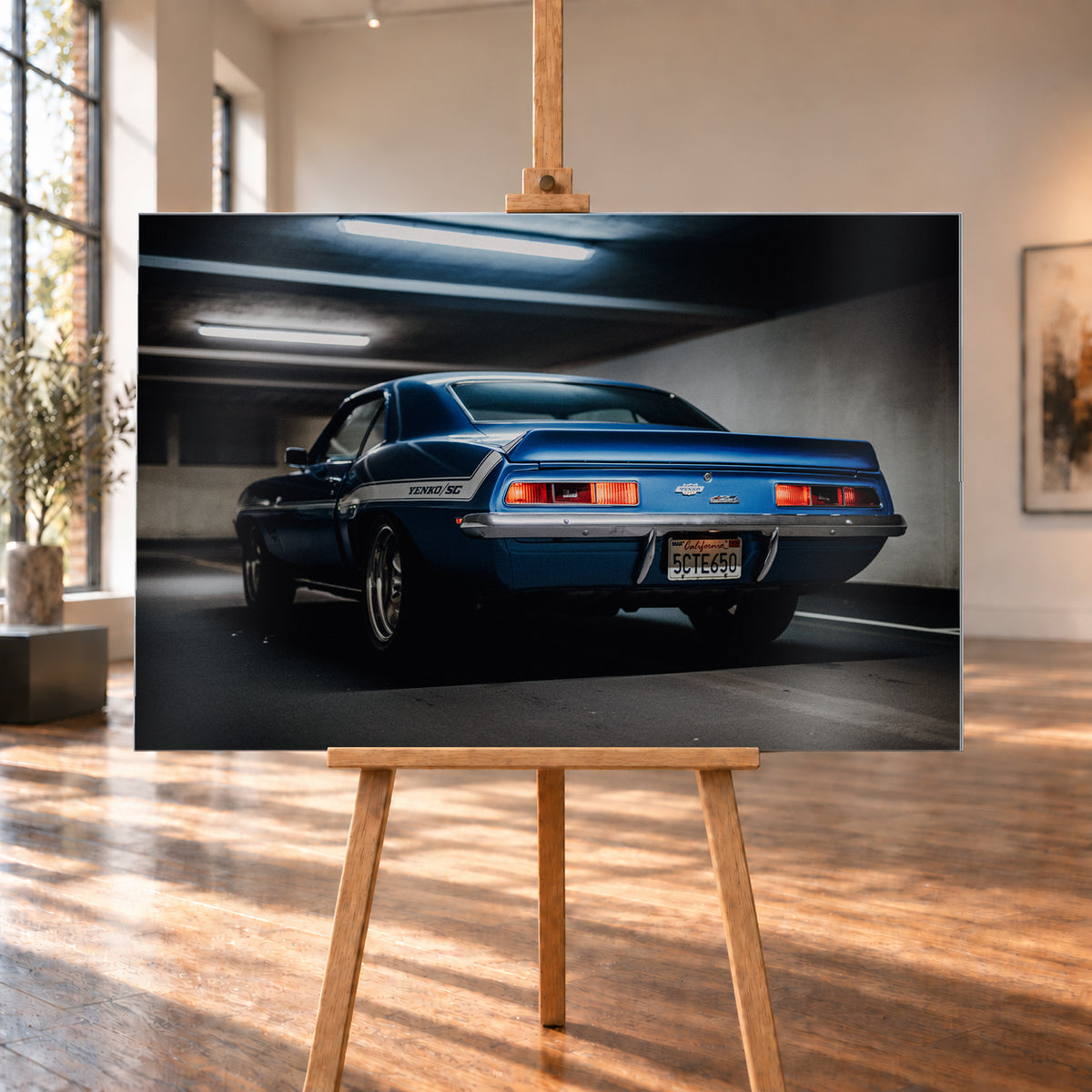 Blauer Chevrolet Camaro Yenko in tiefer Heck-Dreiviertelansicht in einer dunklen Parkgarage; rote Rücklichter, Heckspoiler, YENKO/SC-Seitenstreifen und kühle Deckenleuchten sind sichtbar; Visualisierung als Alu gebürstet-Wandbild, auf einer Holzstaffelei in einem hellen Galerieraum.