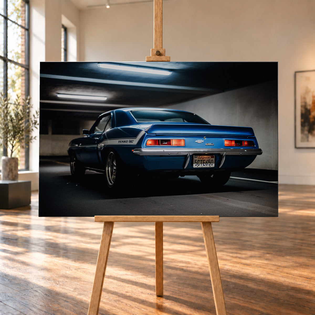 Blauer Chevrolet Camaro Yenko in tiefer Heck-Dreiviertelansicht in einer dunklen Parkgarage; rote Rücklichter, Heckspoiler, YENKO/SC-Seitenstreifen und kühle Deckenleuchten sind sichtbar; Visualisierung als Leinwand-Wandbild, auf einer Holzstaffelei in einem hellen Galerieraum.