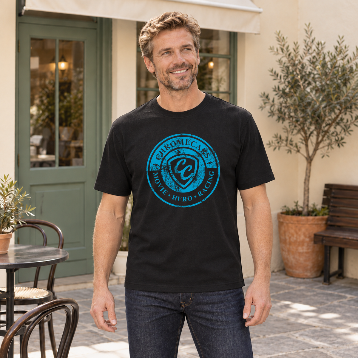 Ein Mann steht lächelnd mit gesenkten Armen im Café-Innenhof und blickt seitlich; sein schwarzes T-Shirt trägt das blaue ChromeCars-Logo vorne.
