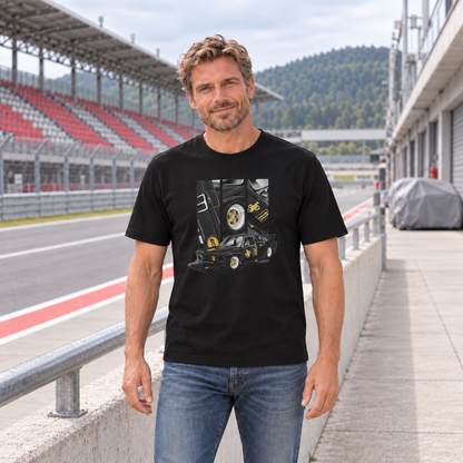 Mann von vorn im schwarzen T-Shirt mit BMW-E21-Motiv auf der Vorderseite, an einer Betonbrüstung vor einer rot-grauen Rennstreckentribüne.