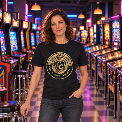 Eine Frau steht in der Retro-Spielhalle, eine Hand am Hocker und eine in der Hosentasche; schwarzes T-Shirt mit goldfarbenem ChromeCars-Logo vorne.