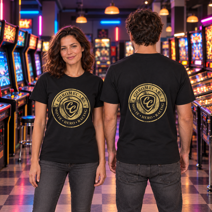 Zwei Personen stehen mit gesenkten Armen in der Retro-Spielhalle: die Frau frontal, der Mann mit dem Rücken zur Kamera; schwarze T-Shirts mit goldfarbenem Logo bei ihr vorne und bei ihm hinten.