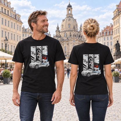 Mann von vorn und Frau von hinten in schwarzen T-Shirts mit Jetta-Motiven, auf einem gepflasterten Stadtplatz vor historischen Gebäuden und einer Kuppelkirche.