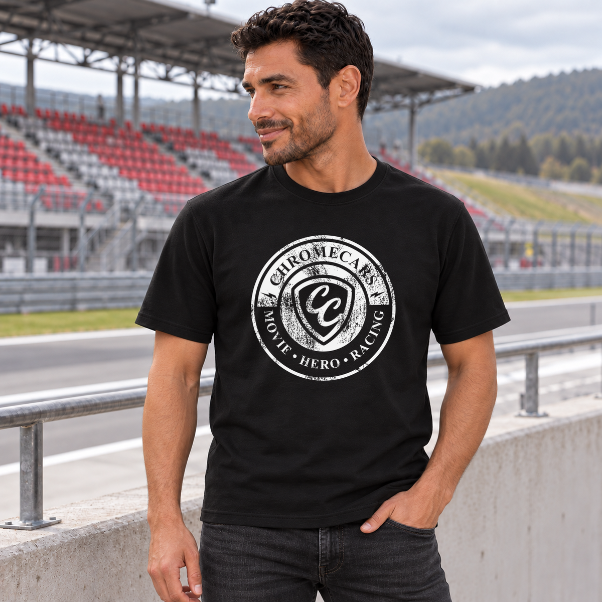 Ein Mann im schwarzen T-Shirt mit weißem Used Logo vorne steht mit einer Hand in der Hosentasche und blickt zur Seite. Hinter der Betonbrüstung liegt die Rennstrecke mit rot-grauer Tribüne.