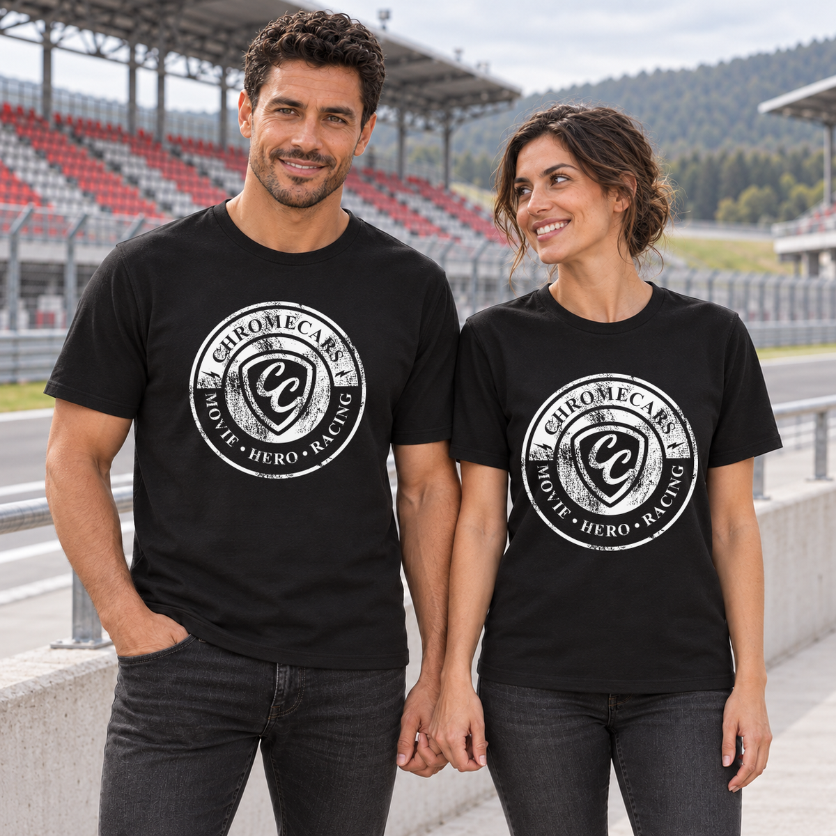 Zwei Personen, ein Mann und eine Frau, stehen händchenhaltend in schwarzen T-Shirts mit weißen Used Logos vorne. Die Frau blickt zum Mann; hinter der Betonbrüstung der Rennstrecke steht eine rot-graue Tribüne.
