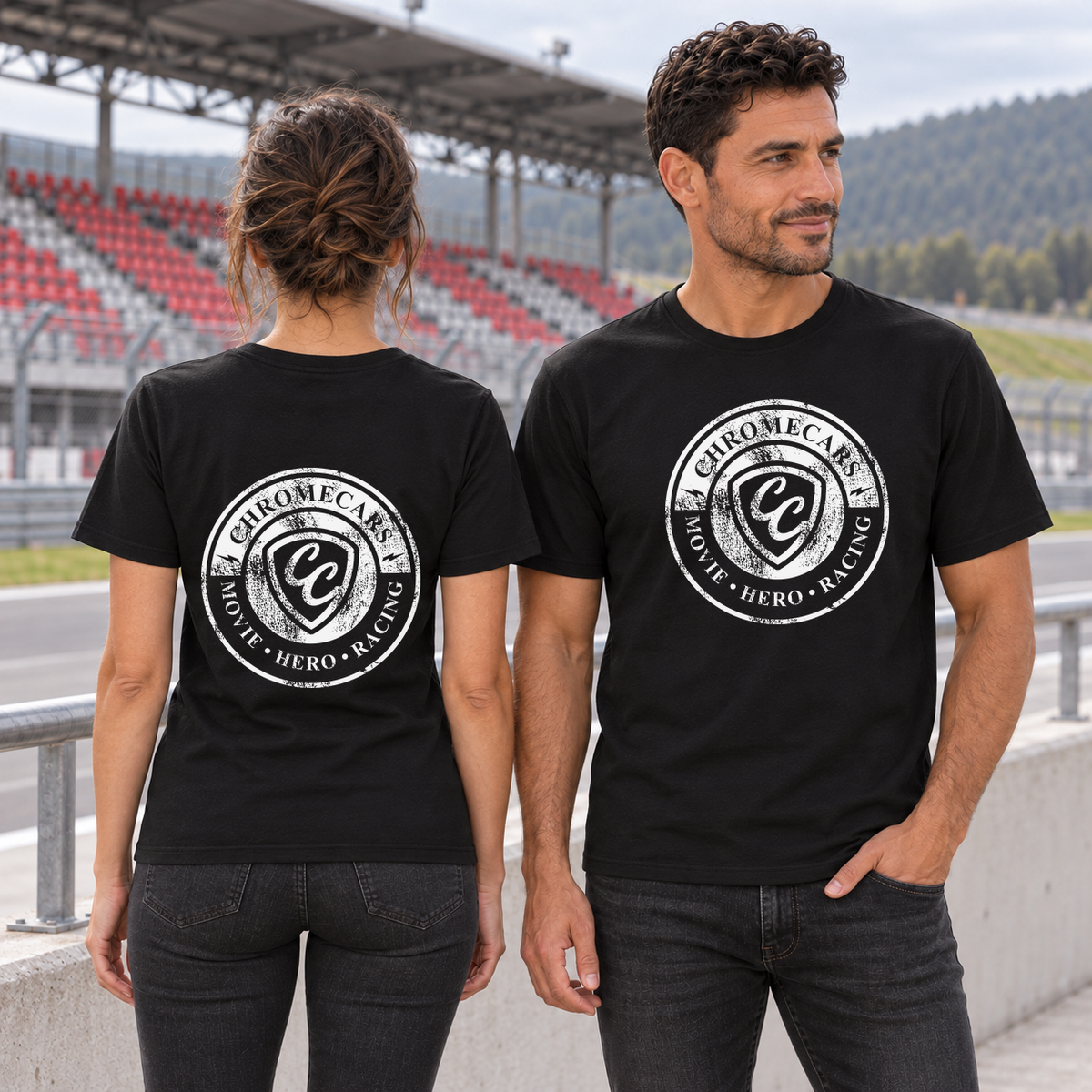 Zwei Personen in schwarzen T-Shirts: links eine Frau mit weißem Used Logo hinten, rechts ein Mann mit weißem Logo vorne und einer Hand in der Hosentasche. Sie stehen an der Rennstrecke vor der rot-grauen Tribüne.