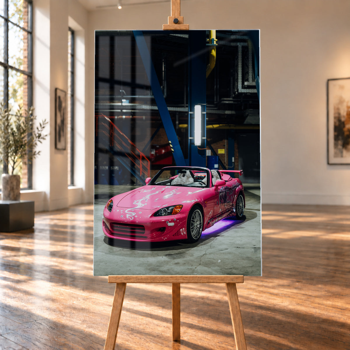 Raumvisualisierung eines hochformatigen Acrylglasbilds auf einer Holzstaffelei zwischen großen Fenstern und hellem Holzboden. Auf der Bildfläche sind Fensterreflexionen sichtbar; das Motiv zeigt den pinken Honda S2000 aus „2 Fast 2 Furious“ in einer dunklen Industriehalle.