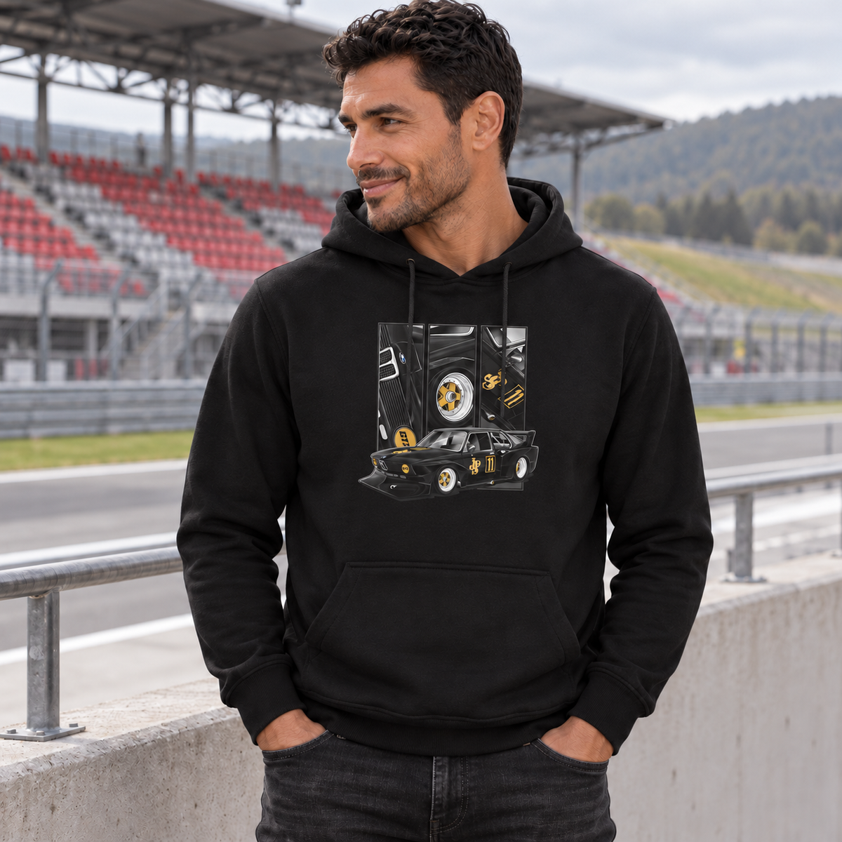 Mann im schwarzen Hoodie lehnt mit einer Hand in der Hosentasche am Geländer einer Rennstreckentribüne. Vorn zeigt der Hoodie einen schwarzgoldenen Rennwagen unter drei Detailfeldern.