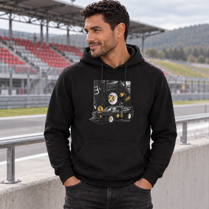 Mann im schwarzen Hoodie lehnt mit einer Hand in der Hosentasche am Geländer einer Rennstreckentribüne. Vorn zeigt der Hoodie einen schwarzgoldenen Rennwagen unter drei Detailfeldern.