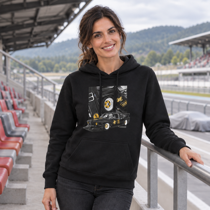 Frau im schwarzen Hoodie steht lächelnd an einem Tribünengeländer vor roten und grauen Sitzen. Das schwarzgoldene Fahrzeugmotiv mit Rad- und Türdetails ist auf ihrer Vorderseite sichtbar.