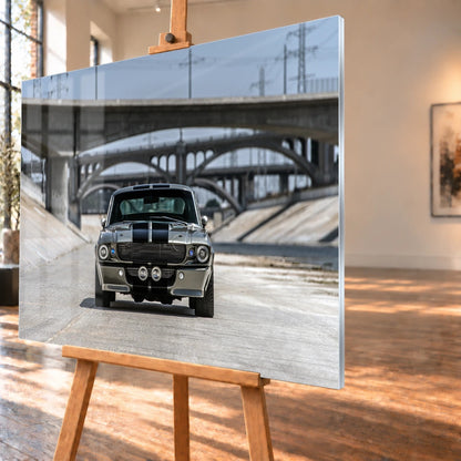 Acrylglas-Visualisierung aus schrägem Blickwinkel mit schmaler heller Seitenkante und Fensterspiegelungen auf der Bildfläche. Das Motiv auf der Holzstaffelei zeigt den Ford Mustang Shelby GT500 „Eleanor“ frontal im Betonkanal unter mehreren Brücken.