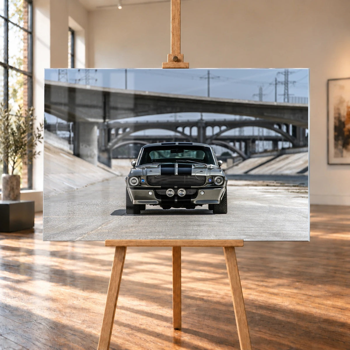 In einem hellen Raum mit hohen Fenstern und Holzboden ist ein Acrylglas-Wandbild auf einer Holzstaffelei visualisiert. Zu sehen ist der silbergraue Ford Mustang Shelby GT500 „Eleanor“ mit schwarzen Doppelstreifen frontal unter Betonbrücken.