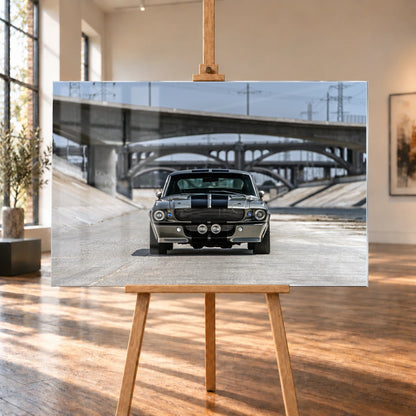 In einem hellen Raum mit hohen Fenstern und Holzboden ist ein Acrylglas-Wandbild auf einer Holzstaffelei visualisiert. Zu sehen ist der silbergraue Ford Mustang Shelby GT500 „Eleanor“ mit schwarzen Doppelstreifen frontal unter Betonbrücken.