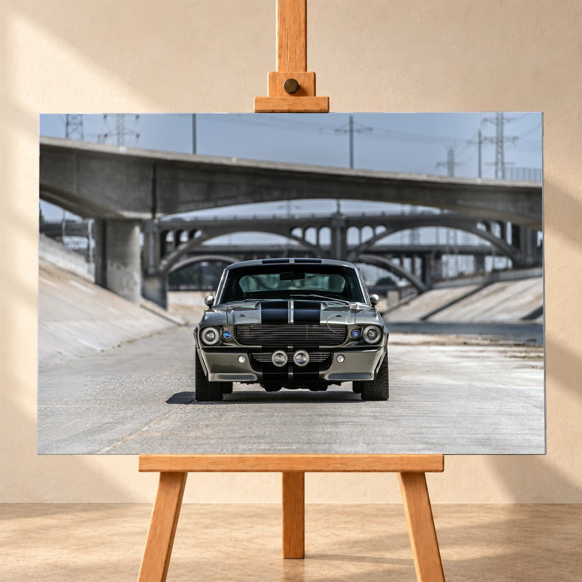 Frontale Visualisierung eines Wandbilds aus gebürstetem Aluminium auf einer Holzstaffelei vor beiger Wand. Das querformatige Motiv zeigt den Ford Mustang Shelby GT500 „Eleanor“ mit schwarzen Doppelstreifen in einem Betonkanal vor mehreren Bogenbrücken.