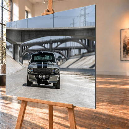 Schräg visualisiertes Wandbild aus gebürstetem Aluminium auf einer Holzstaffelei; rechts ist die dünne dunkle Plattenkante sichtbar. Im Motiv steht der silbergraue Ford Mustang Shelby GT500 „Eleanor“ frontal im Betonkanal unter gestaffelten Brücken.