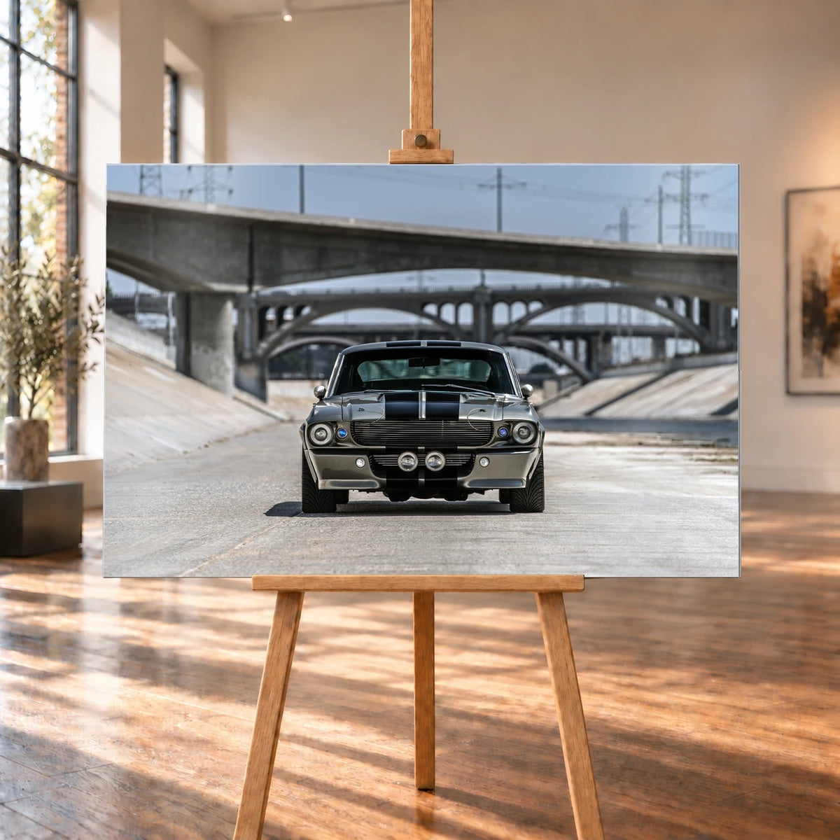Raumansicht als Visualisierung: Ein querformatiges Wandbild aus gebürstetem Aluminium steht auf einer Holzstaffelei zwischen hohen Fenstern und sonnenbeschienenem Holzboden. Es zeigt den Ford Mustang Shelby GT500 „Eleanor“ frontal vor Betonbrücken.