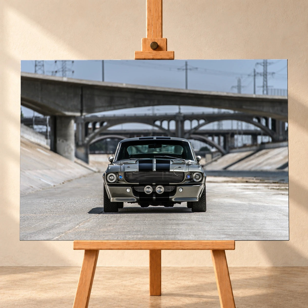 Visualisierung einer Leinwand im Querformat auf einer Holzstaffelei vor heller Wand. Das Motiv zeigt den silbergrauen Ford Mustang Shelby GT500 „Eleanor“ mit schwarzen Doppelstreifen frontal in einem Betonkanal unter mehreren Brücken.