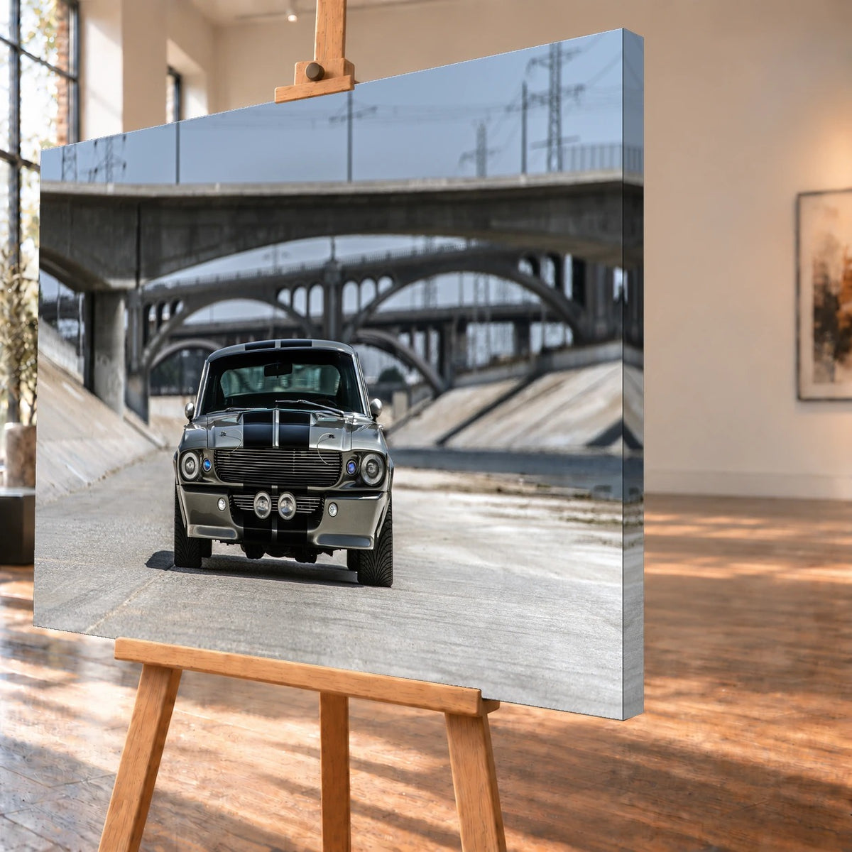 Schrägansicht einer visualisierten Leinwand auf einer Holzstaffelei, mit sichtbarer bedruckter rechter Seitenkante. Auf dem Motiv steht der silbergraue Ford Mustang Shelby GT500 „Eleanor“ mit schwarzen Doppelstreifen frontal vor gestaffelten Betonbrücken.