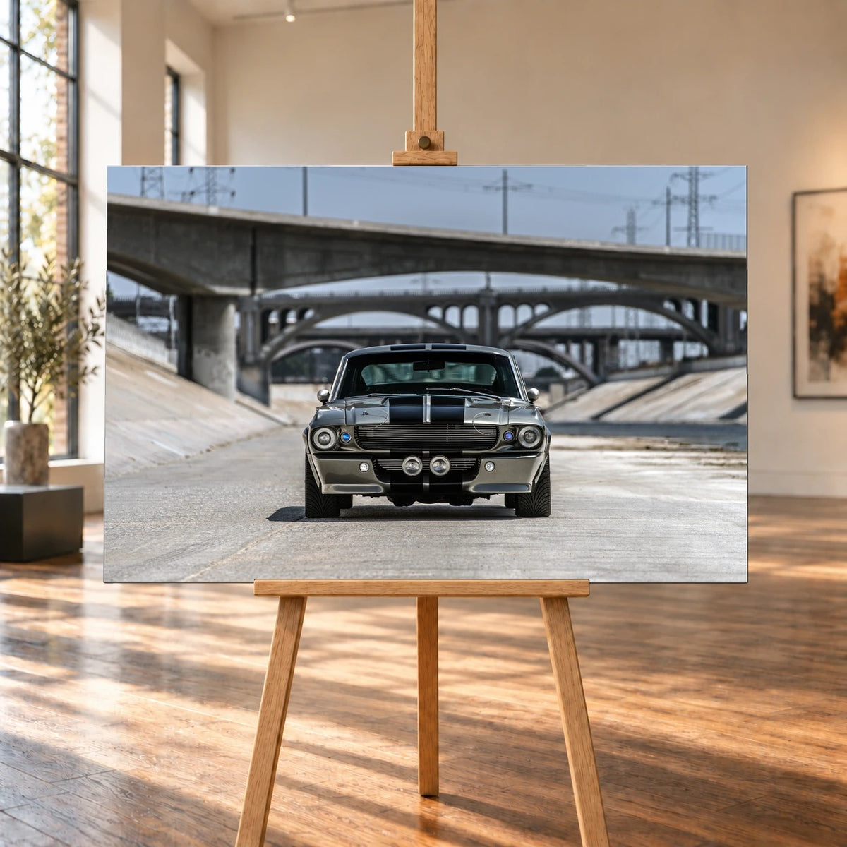 Raumvisualisierung mit einer querformatigen Leinwand auf einer Holzstaffelei, umgeben von hohen Fenstern und sonnenbeschienenem Holzboden. Das Bild zeigt den Ford Mustang Shelby GT500 „Eleanor“ in Frontalansicht im Betonkanal unter Bogenbrücken.