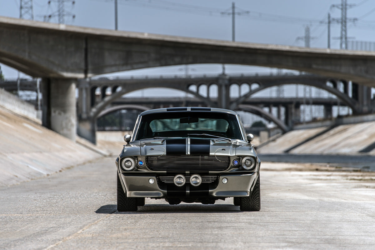 Silbergrauer Ford Mustang Shelby GT500 „Eleanor“ mit schwarzen Doppelstreifen und runden Zusatzscheinwerfern in Frontalansicht in einem Betonkanal. Hinter dem Wagen staffeln sich mehrere Bogenbrücken, darüber verlaufen Stromleitungen.