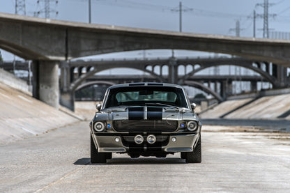 Silbergrauer Ford Mustang Shelby GT500 „Eleanor“ mit schwarzen Doppelstreifen und runden Zusatzscheinwerfern in Frontalansicht in einem Betonkanal. Hinter dem Wagen staffeln sich mehrere Bogenbrücken, darüber verlaufen Stromleitungen.