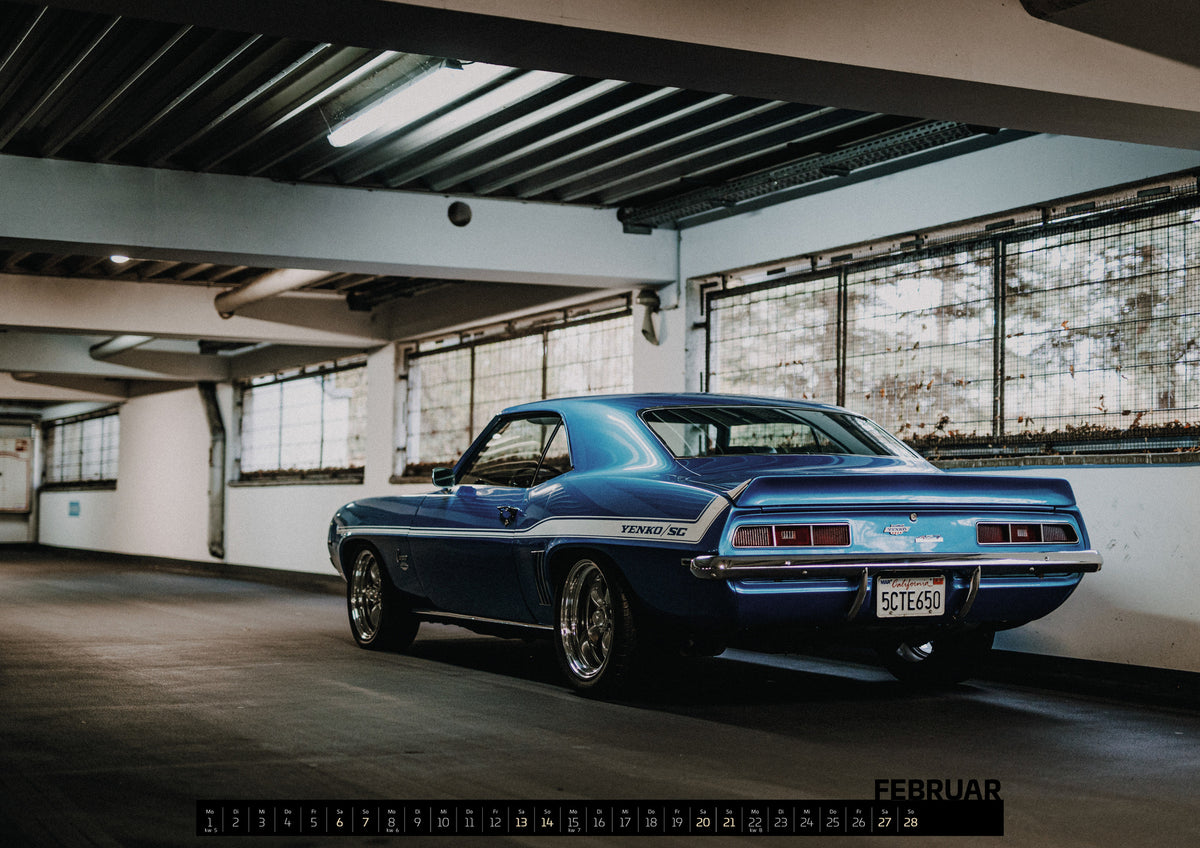 Yenko Camaro Kalender 2027 – Februar: Der Camaro Yenko steht schräg von hinten in einer weiten Industriehalle; Heck, Spoiler und weißer Seitenstreifen heben sich vom Beton ab