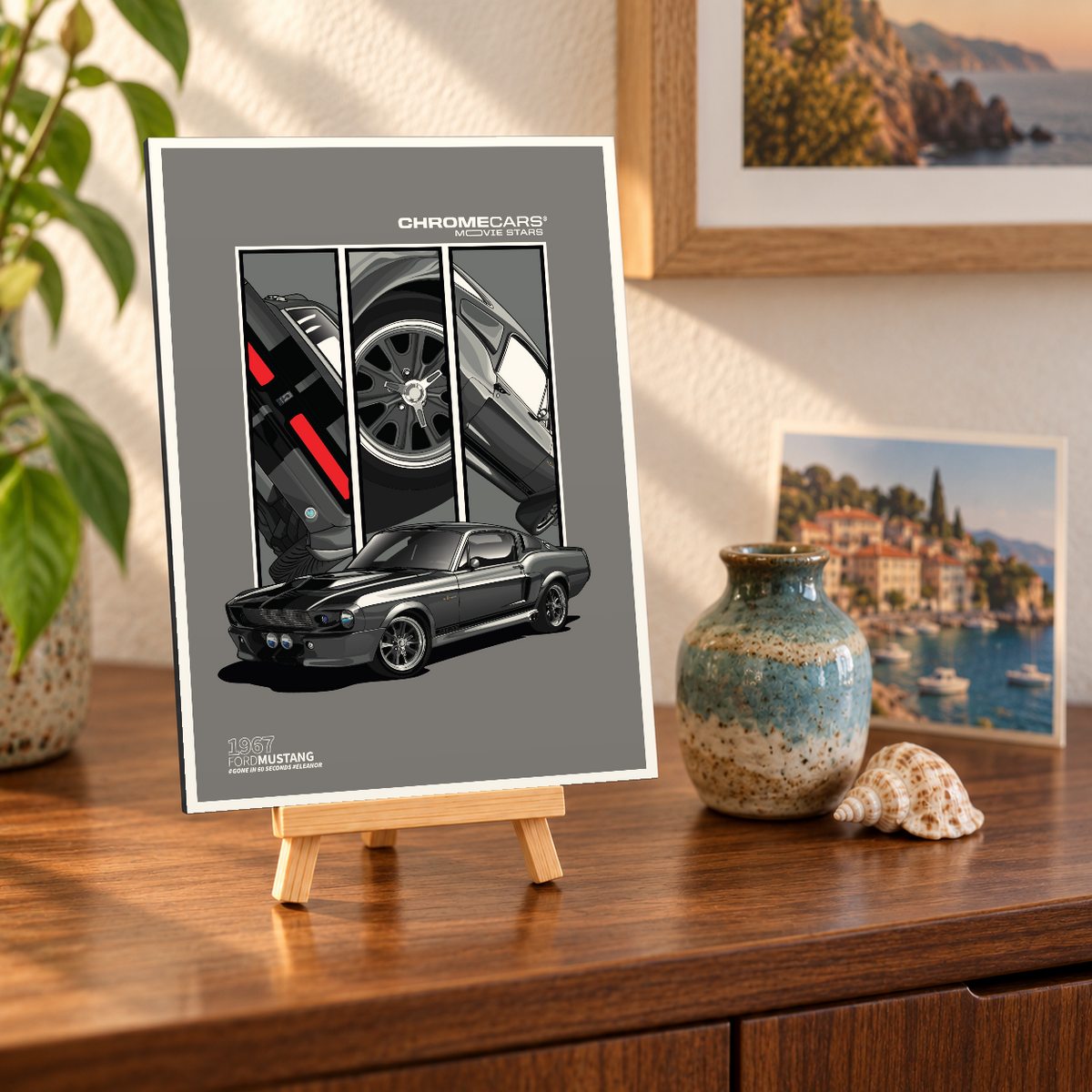 Mockup einer kleinen hochformatigen Alu-Dibond-Bildplatte auf einer Holzstaffelei, leicht schräg von vorn auf einer Holzkommode gezeigt. Das Motiv zeigt den dunkelgrauen Ford Mustang „ELEANOR“ in vorderer Dreiviertelansicht vor drei Detailfeldern mit Heckleuchten, Rad und Karosserie. Vase, Muschel, Pflanzen und Küstenbilder gehören zur Szenerie, nicht zum Lieferumfang.