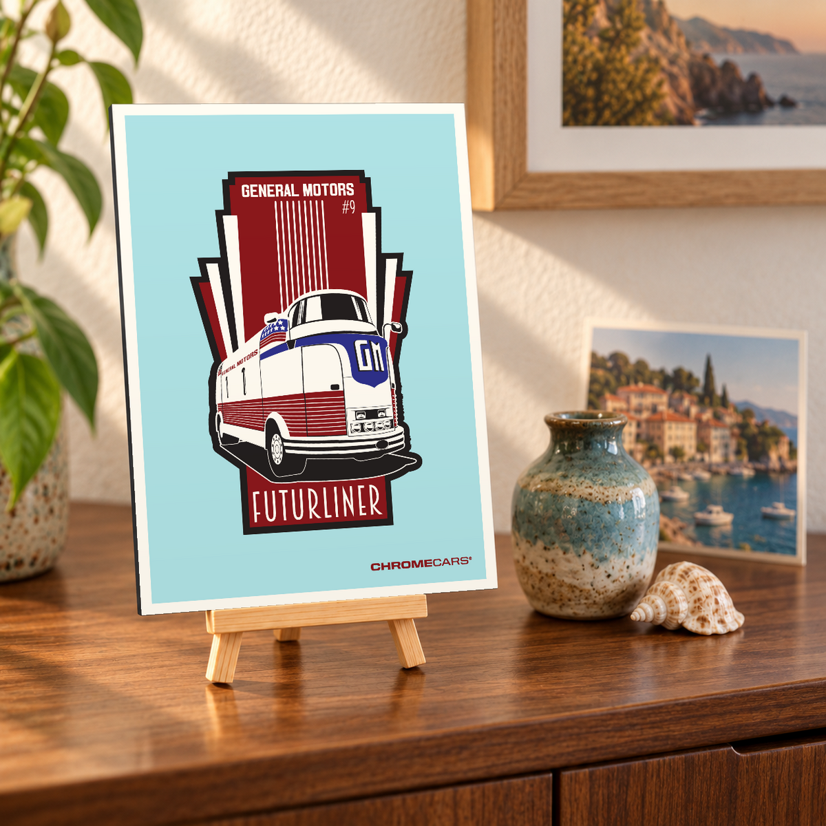 Mockup einer kleinen Alu-Dibond-Bildplatte im Hochformat auf einer Holzstaffelei, leicht schräg auf einer Holzkommode betrachtet. Das Motiv zeigt den rot-weißen GM Futurliner #9 mit blauer Frontpartie in Dreiviertelansicht vor türkisfarbenem Hintergrund und rot-schwarzer Grafik. Pflanze, Vase, Muschel und Küstenbilder bilden die Szenerie und gehören nicht zum Lieferumfang.