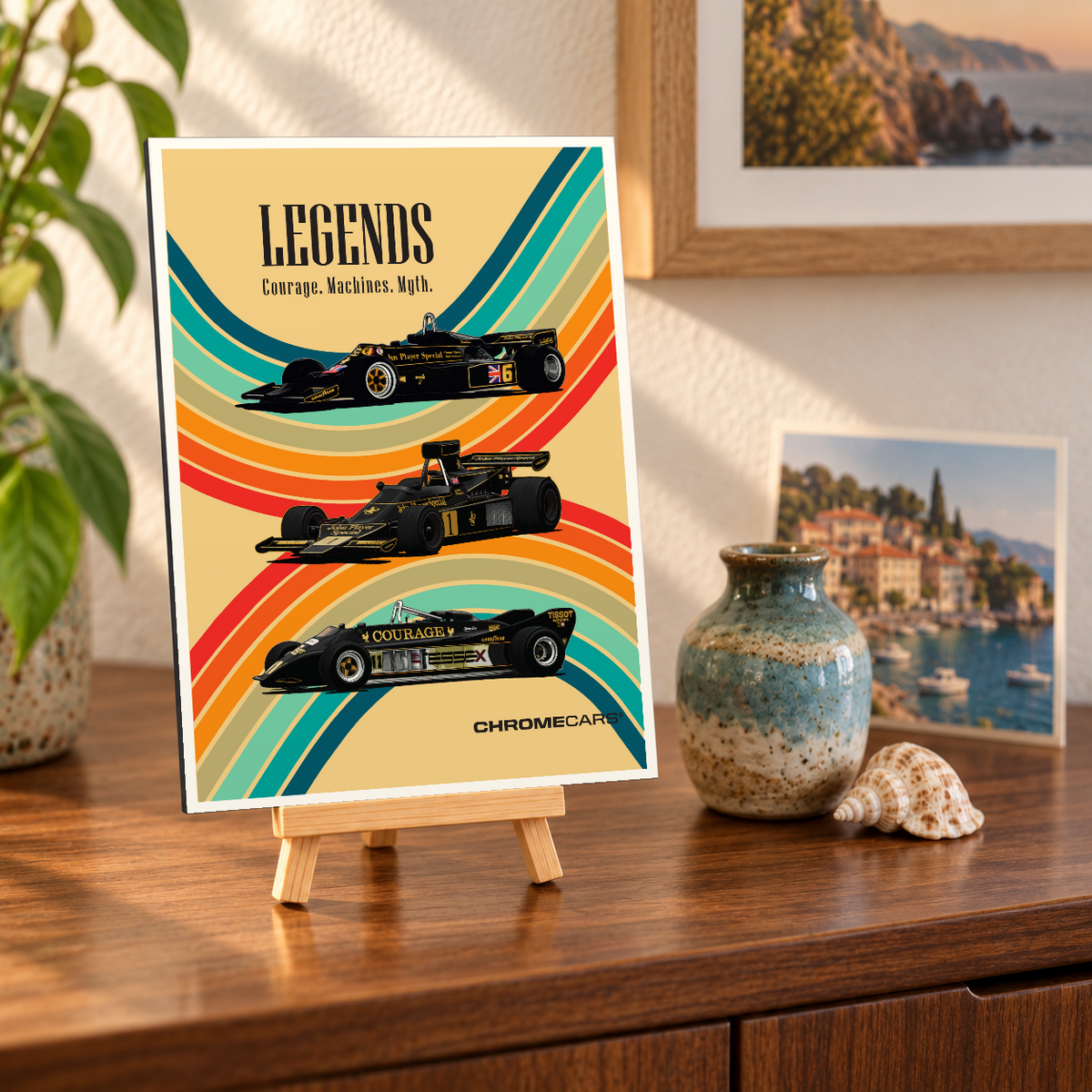 Mockup einer kleinen hochformatigen Alu-Dibond-Bildplatte auf einer Holzstaffelei, leicht schräg von vorn auf einer Holzkommode gezeigt. Das Motiv zeigt drei schwarz-goldene Formelrennwagen vor geschwungenen bunten Streifen auf beigefarbenem Grund und die Überschrift „LEGENDS“. Pflanze, Vase, Muschel und Küstenbilder sind Teil der Szenerie, nicht des Lieferumfangs.