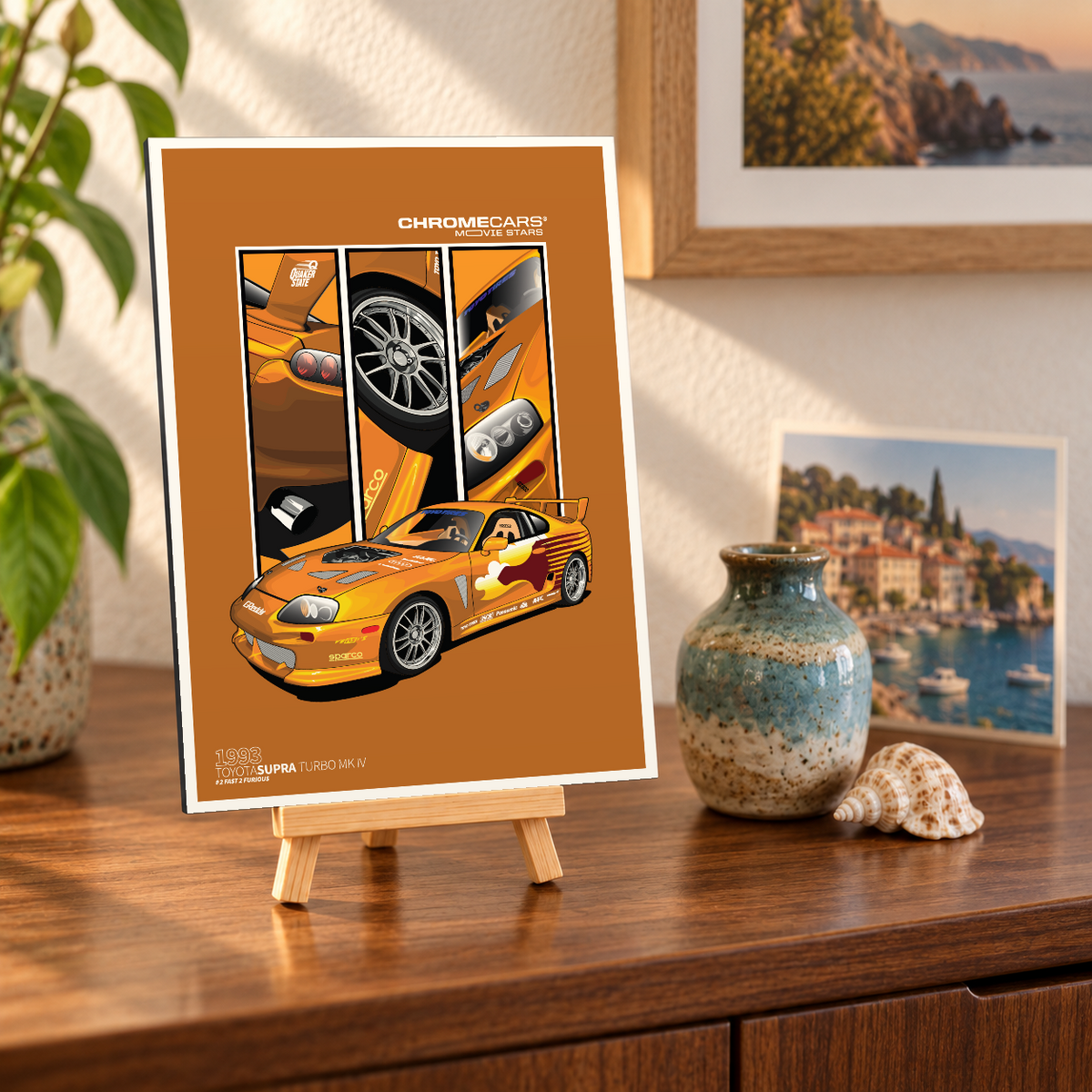 Mockup einer kleinen hochformatigen Alu-Dibond-Bildplatte auf einer Holzstaffelei, leicht schräg auf einer dunklen Holzkommode gezeigt. Das Motiv zeigt den orangefarbenen Fast 2 „Slap Jack“ Supra in vorderer Dreiviertelansicht vor drei senkrechten Detailbildern von Heck, Rad und Front auf braunorangefarbenem Grund. Vase, Muschel, Pflanze und Küstenbilder bilden die Szenerie.