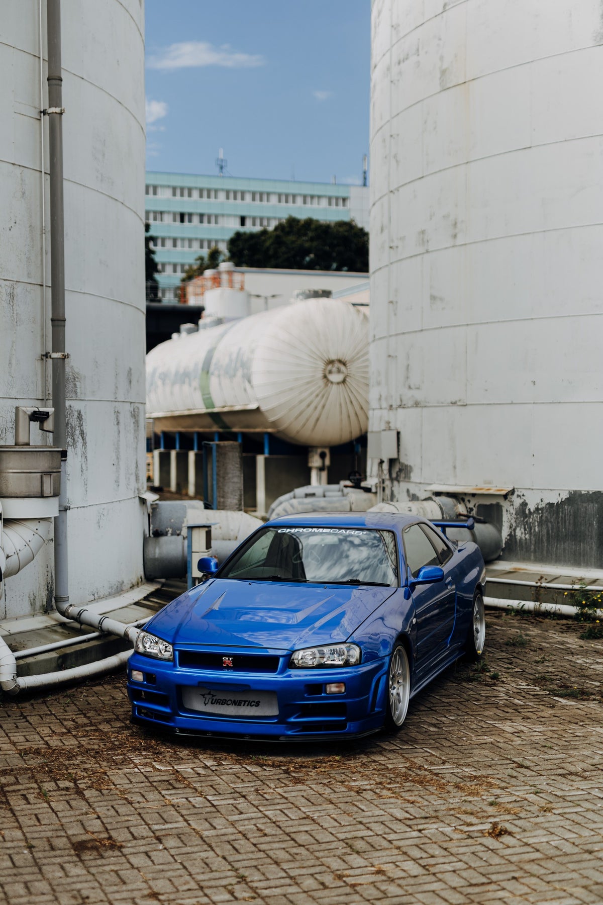 Blauer Nissan Skyline R34 GT-R in frontaler Dreiviertelansicht auf gepflastertem Industriegelände zwischen hohen weißen Behältern, Tanks und Rohrleitungen bei Tageslicht.