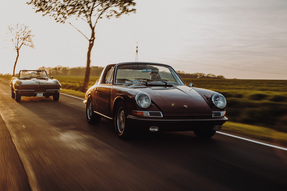 Ein dunkelrot wirkender Porsche 911 S Softwindow fährt rechts im Vordergrund, dahinter links eine dunkle Chevrolet Corvette C2; beide mit offenem Dach und sichtbaren Fahrern auf einer Landstraße bei tiefem, warmem Gegenlicht.