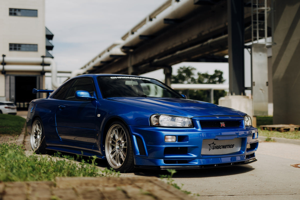 Blauer Nissan Skyline R34 GT-R in tiefer Front-Dreiviertelansicht auf Asphalt unter einer erhöhten Betonstruktur; sichtbar sind silberne Mehrspeichenräder, Heckflügel, GT-R-Emblem und Gras am Bildrand.