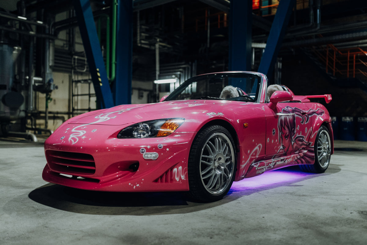 Pinker Honda S2000 als offener Roadster in Front-Dreiviertelansicht auf einem Betonboden in einer dunklen Industriehalle; sichtbar sind weiße Dekorgrafiken, eine große Frauenfigur an der Seite, silberne Mehrspeichenräder, ein Heckflügel und violett-blaues Licht unter dem Fahrzeug.
