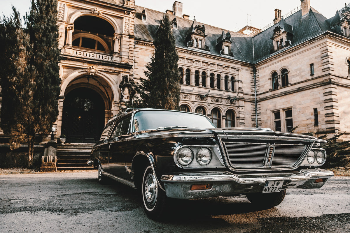 Dunkler Chrysler New Yorker Town & Country in niedriger Front-Dreiviertelansicht vor einem großen historischen Steingebäude mit Rundbögen; warmes Tageslicht spiegelt sich in Chromfront und Lack.