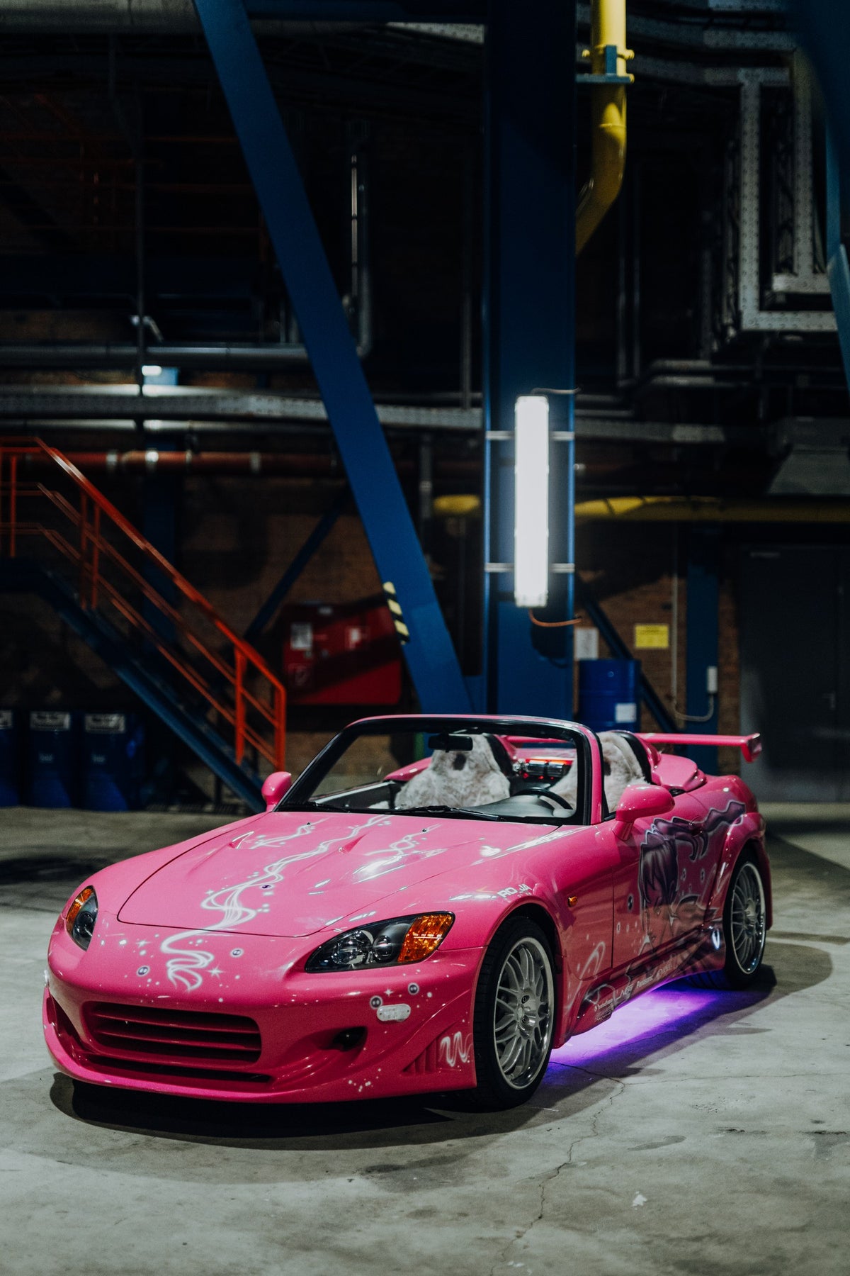 Der pinke Honda S2000 aus „2 Fast 2 Furious“ steht mit offenem Verdeck schräg von vorn in einer dunklen Industriehalle. Weißes Dekor und eine große Seitengrafik zieren die Karosserie; unter dem Wagen leuchtet violettes Licht.