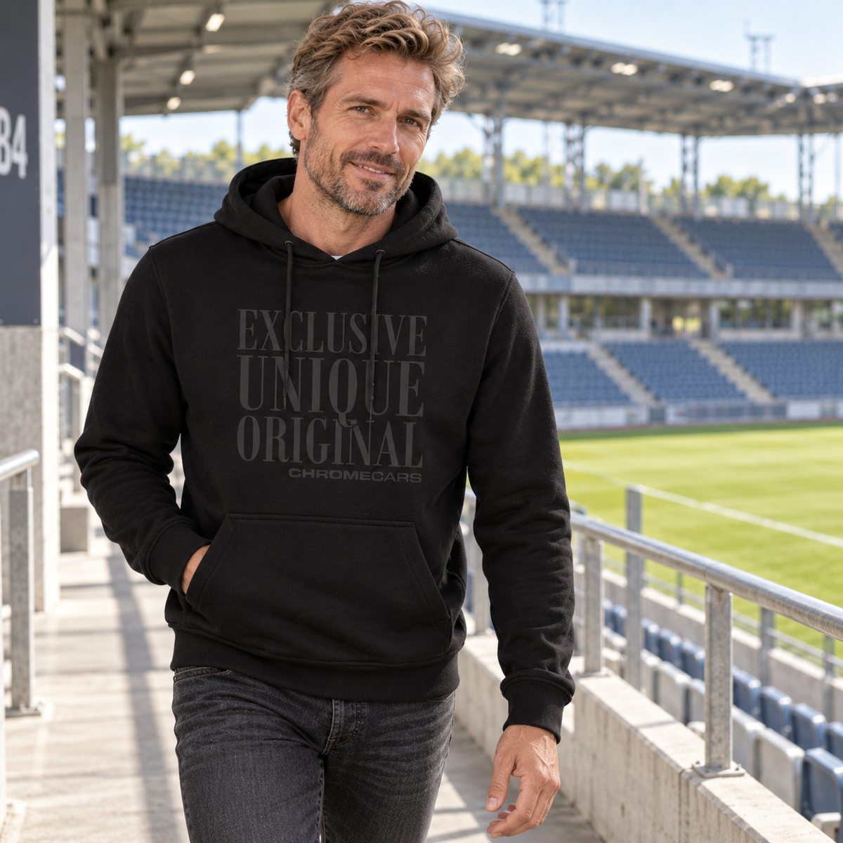 Ein Mann im schwarzen Hoodie mit dezentem schwarzem Typografiemotiv auf einem Tribünengang im Fußballstadion; eine Hand steckt in der Kängurutasche, der andere Arm hängt seitlich.